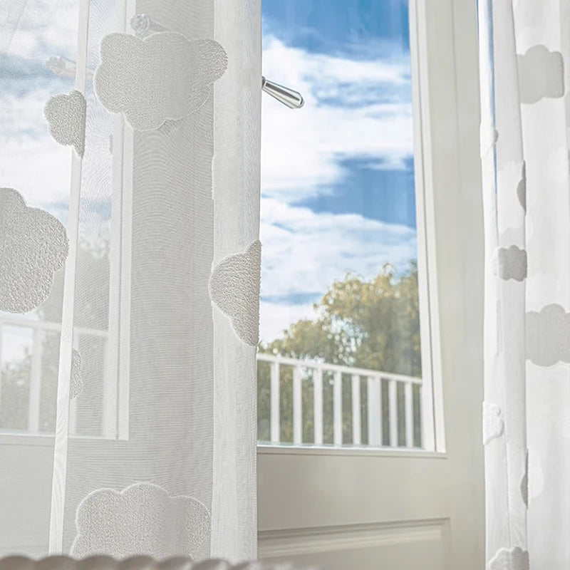 Voilage blanc à motifs de nuages devant une porte-fenêtre ouverte sur un ciel bleu, vue sur un balcon arboré.