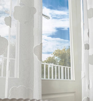 Voilage blanc à motifs de nuages devant une porte-fenêtre ouverte sur un ciel bleu, vue sur un balcon arboré.