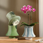 Une magnifique orchidée aux fleurs rose vif trône sur une table en bois, entourée de vases vintage en céramique verte et bleu clair.