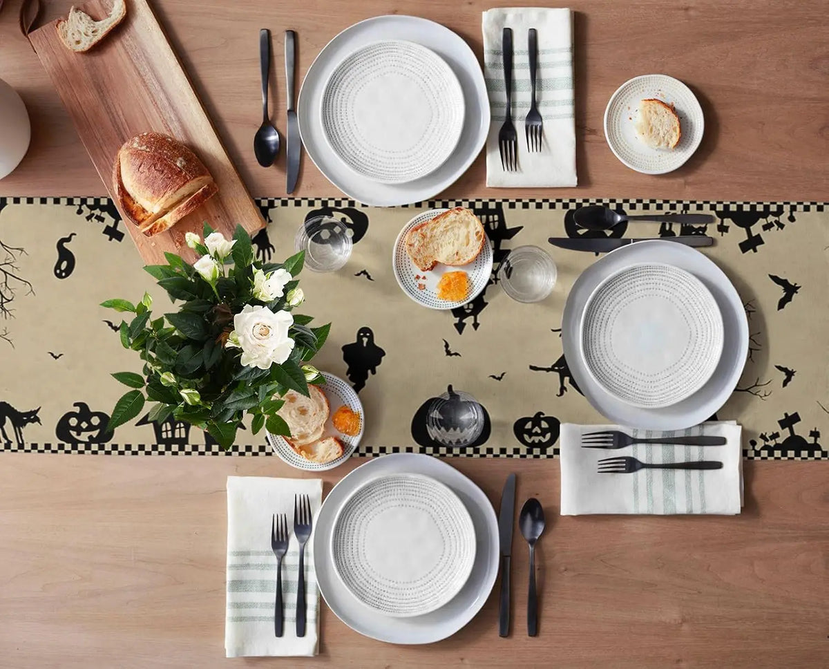 La table est joliment décorée avec des assiettes blanches, des couverts noirs, des pains ronds, des fleurs blanches et des accessoires Halloween. Une mise en scène élégante et festive pour un repas d'automne.