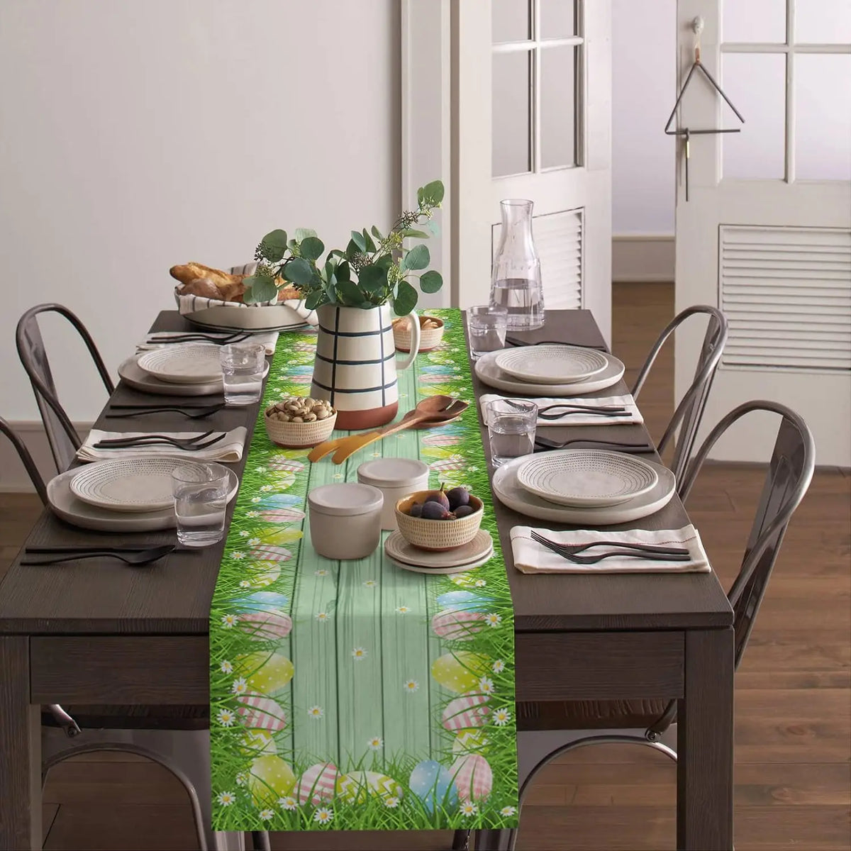 Une table dressée pour un repas, avec une nappe ornée de motifs floraux et de couleurs printanières, des assiettes blanches, des verres et des accessoires de table.