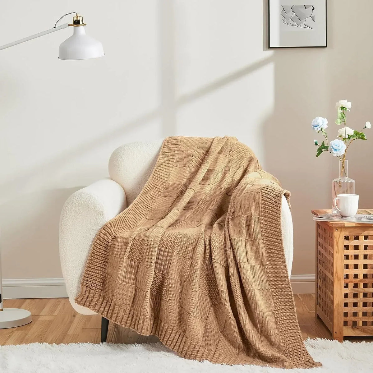 Une couverture en tricot beige posée sur un fauteuil douillet dans un intérieur cosy, avec une lampe blanche et un bouquet de fleurs bleues sur une table basse en bois.