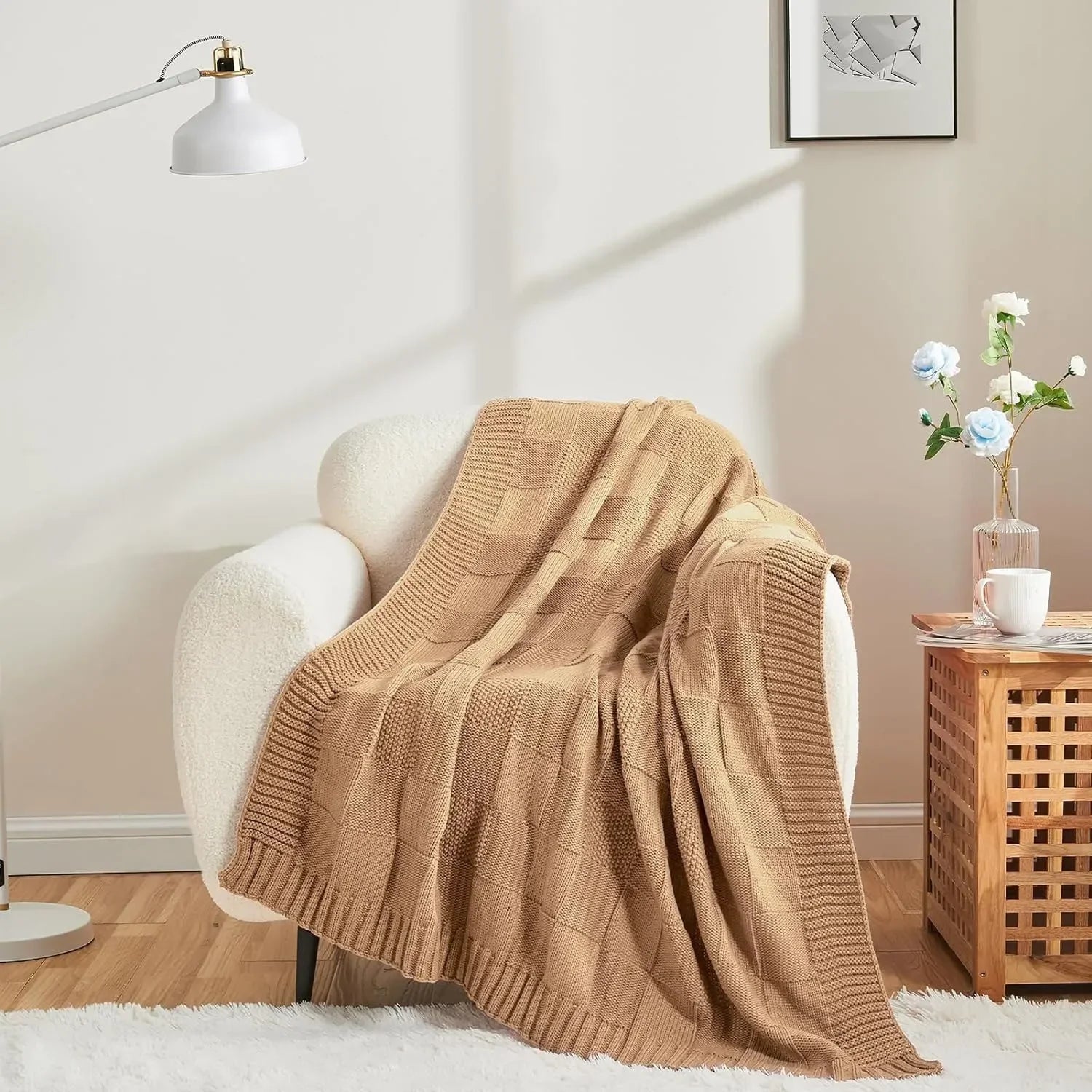 Une couverture en tricot beige posée sur un fauteuil douillet dans un intérieur cosy, avec une lampe blanche et un bouquet de fleurs bleues sur une table basse en bois.