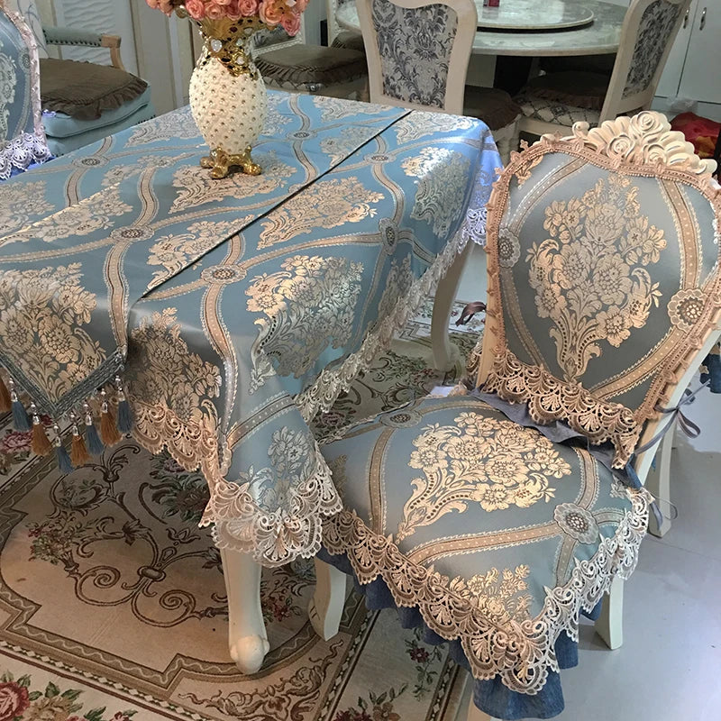 Cette image montre une table ronde recouverte d'une nappe en tissu orné de motifs floraux dorés sur un fond bleu pâle, entourée de chaises aux dossiers sculptés et décorés de dentelle.