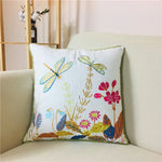 Cette image représente un coussin décoratif orné de motifs floraux et d'insectes, tels que des libellules, dans des couleurs vives et joyeuses, créant une atmosphère chaleureuse et naturelle dans l'espace.