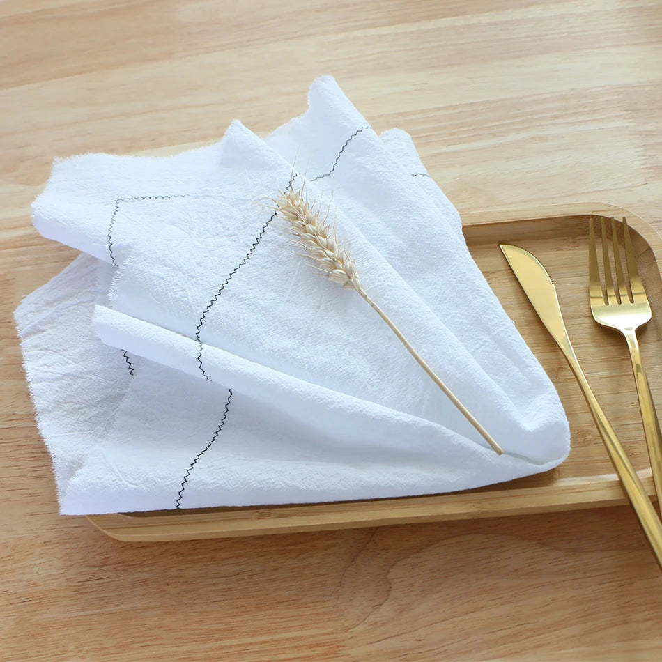Une serviette en tissu blanc posée sur une surface en bois, accompagnée d'une tige de blé et de couverts dorés, créant une atmosphère élégante et raffinée dans un cadre simple et épuré.
