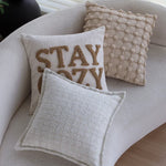 Cette image montre des coussins décoratifs moelleux avec un motif texturé et le mot "STAY AWAY" brodé en lettres dorées, créant une ambiance confortable et accueillante dans un espace de détente.