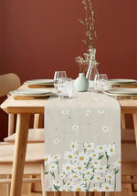 Une table élégante avec une nappe blanche à motifs de marguerites, entourée de vaisselle et de verres sur un fond brun, créant une ambiance chaleureuse.