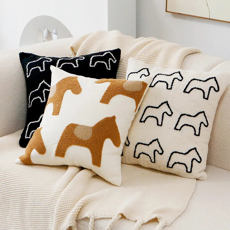 Des coussins décoratifs avec des motifs de chevaux stylisés, posés sur une couverture douillette dans une ambiance chaleureuse et minimaliste.