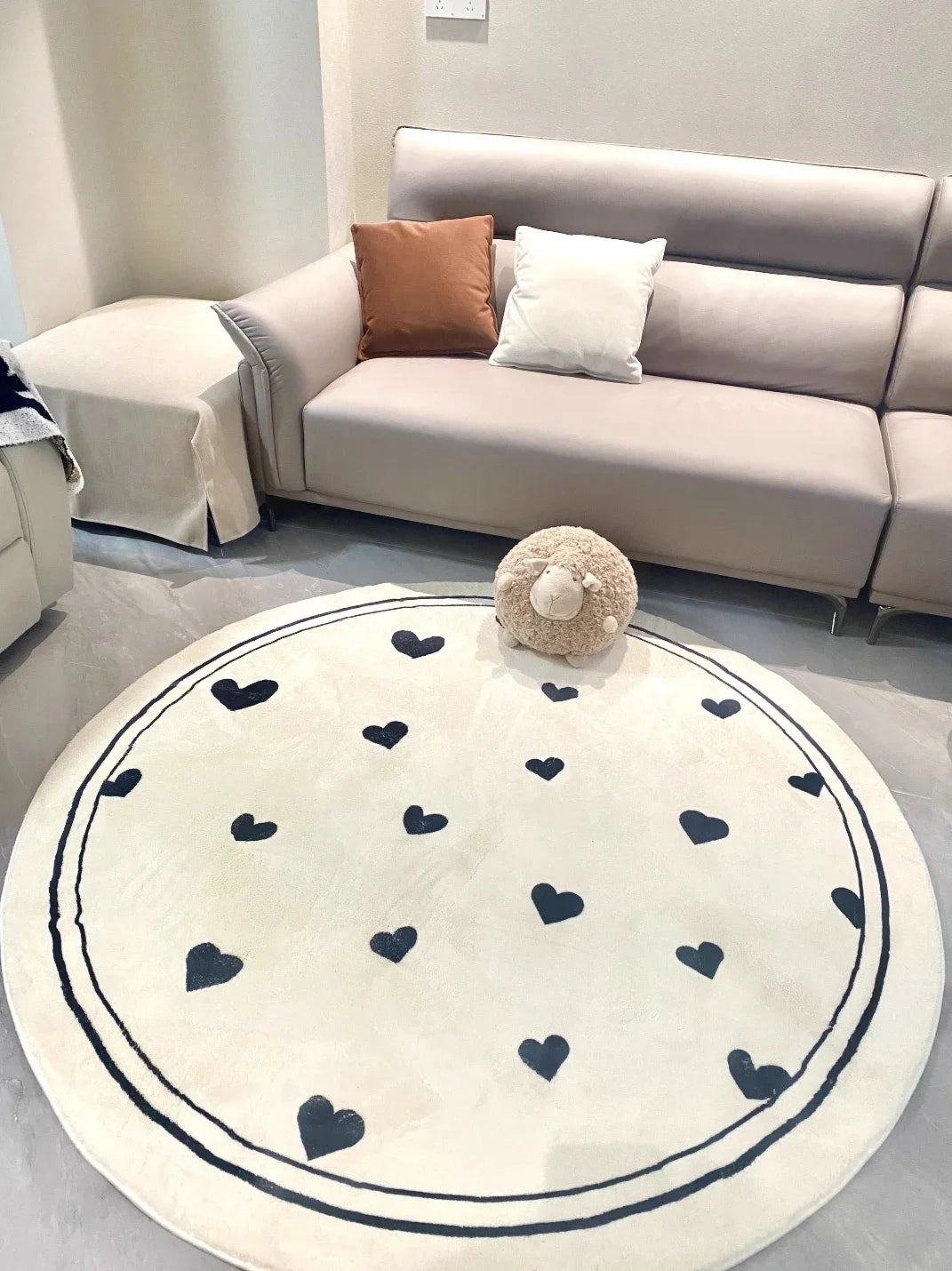 Un tapis rond blanc avec des motifs de coeurs noirs décore un salon cosy avec un canapé gris et des oreillers décoratifs.