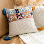 Coussin décoratif avec motifs géométriques colorés et glands bleu et jaune sur fond beige et rayé.