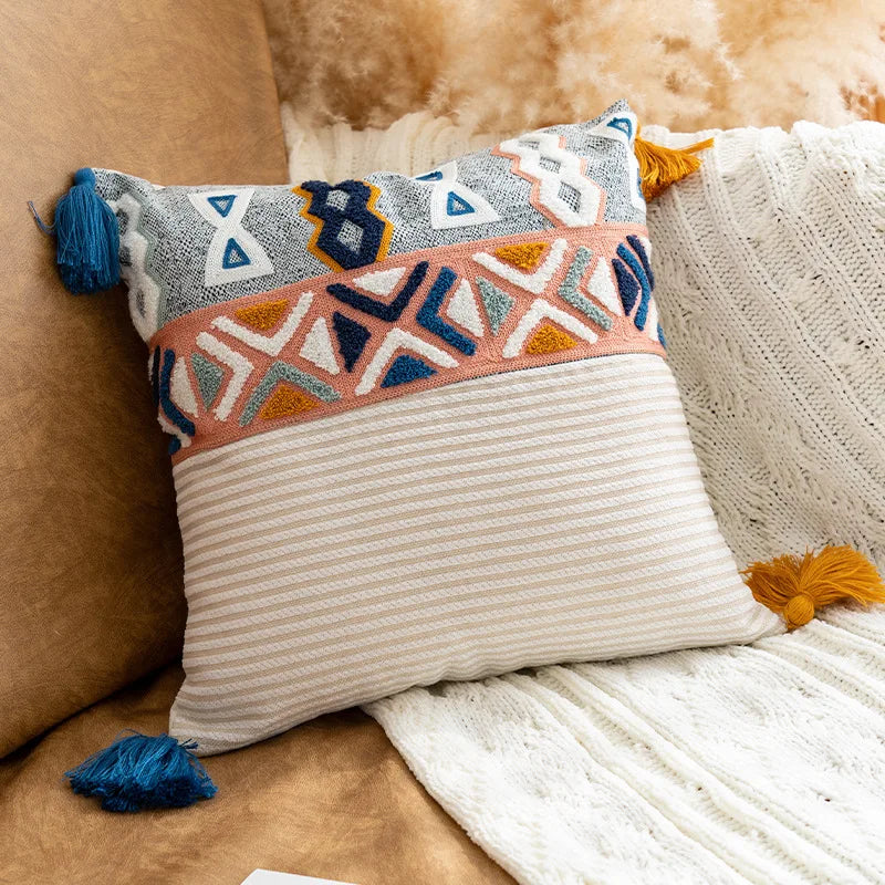 Coussin décoratif avec motifs géométriques colorés et glands bleu et jaune sur fond beige et rayé.