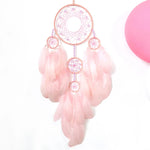 Un capteur de rêves rose avec des plumes et des cercles métalliques suspendus sur un fond blanc.