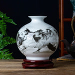 Un vase en porcelaine blanche décorée de motifs floraux en nuances de noir et de blanc, posé sur un support en bois, entouré de plantes vertes.
