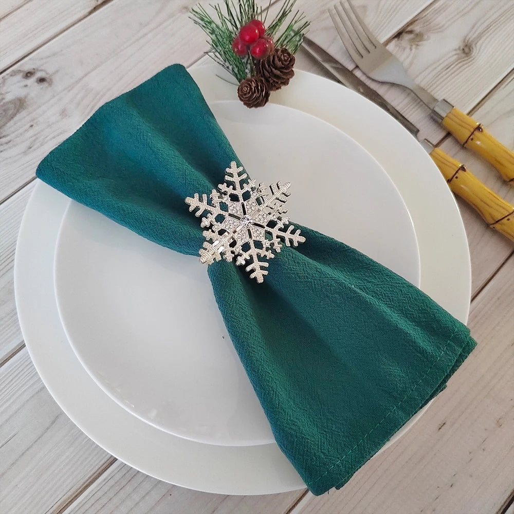 Sur une assiette blanche posée sur un fond en bois clair, une serviette de table verte est pliée et ornée d'un élégant cristal en forme de flocon de neige, avec des cônes de pin et des baies rouges ajoutés comme touche festive.