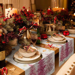 Cette scène de table de Noël chaleureuse et festive met en valeur des décorations rouges et vertes, des bougies allumées, et une belle vaisselle sur un linge de table fleuri, créant une ambiance conviviale et élégante pour les festivités.