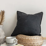 Un coussin décoratif noir posé sur un panier tressé en fibre naturelle, entouré d'accessoires de décoration dans un intérieur moderne et épuré.