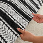 L'image montre un motif noir et blanc complexe et géométrique sur un tapis ou un tissu, avec des mains féminine sur le côté pour apprécier la texture et les couleurs.