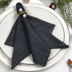 Un élégant ensemble de serviettes noires avec des anneaux dorés et des perles sur une assiette blanche, entouré de fleurs.