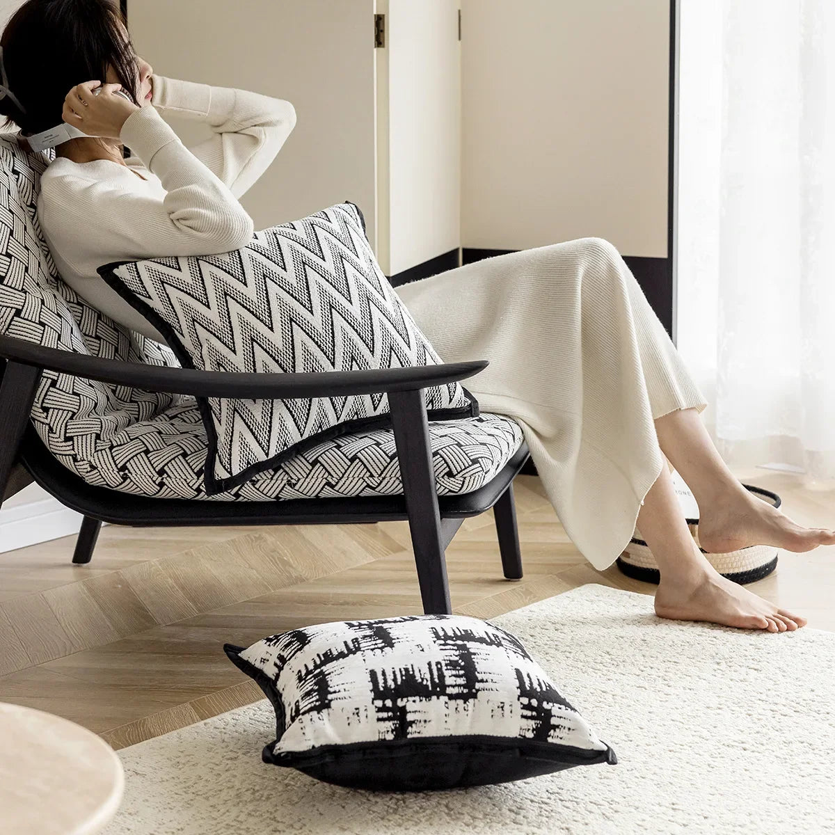 L'image montre une personne assise confortablement dans un fauteuil d'intérieur avec des motifs noirs et blancs. La personne a les pieds nus et est enveloppée dans une couverture blanche. Des coussins décoratifs ornent le fauteuil, créant une atmosphère chaleureuse et apaisante.