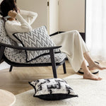 L'image montre une personne assise confortablement dans un fauteuil d'intérieur avec des motifs noirs et blancs. La personne a les pieds nus et est enveloppée dans une couverture blanche. Des coussins décoratifs ornent le fauteuil, créant une atmosphère chaleureuse et apaisante.