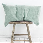 Coussin en lin vert posé sur un tabouret rustique en bois, créant une ambiance sereine et naturelle.
