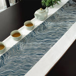 Sur la table, une théière blanche et des tasses remplies de thé sont disposées sur un élégant chemin de table bleu foncé orné de motifs ondulés.