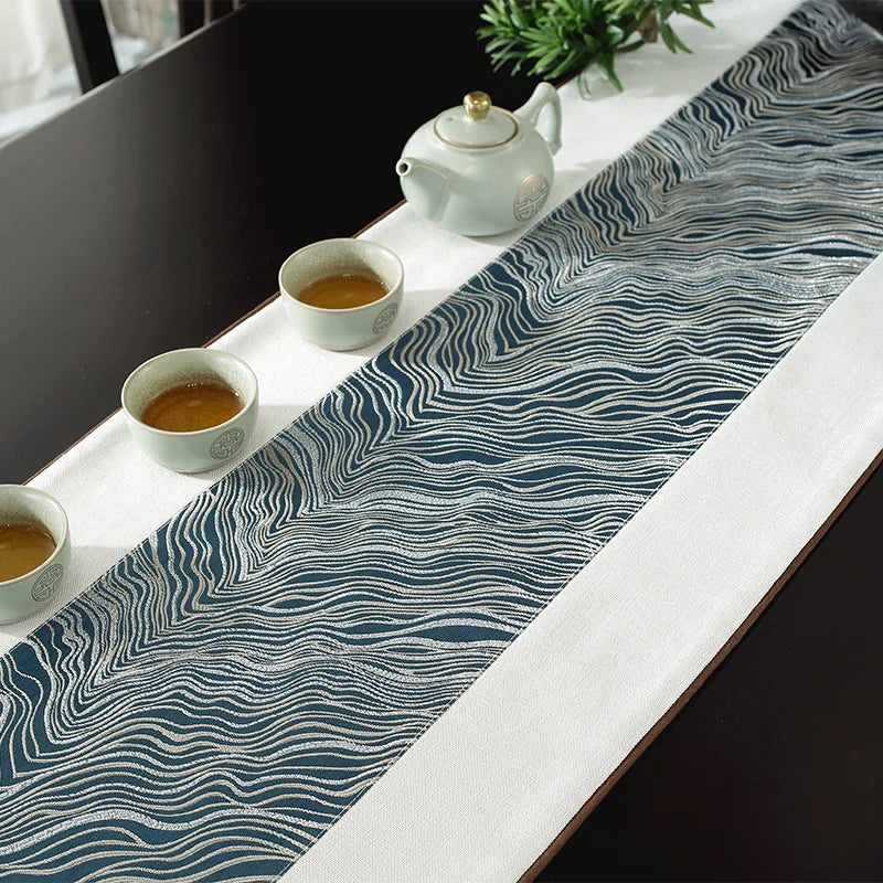 Sur la table, une théière blanche et des tasses remplies de thé sont disposées sur un élégant chemin de table bleu foncé orné de motifs ondulés.