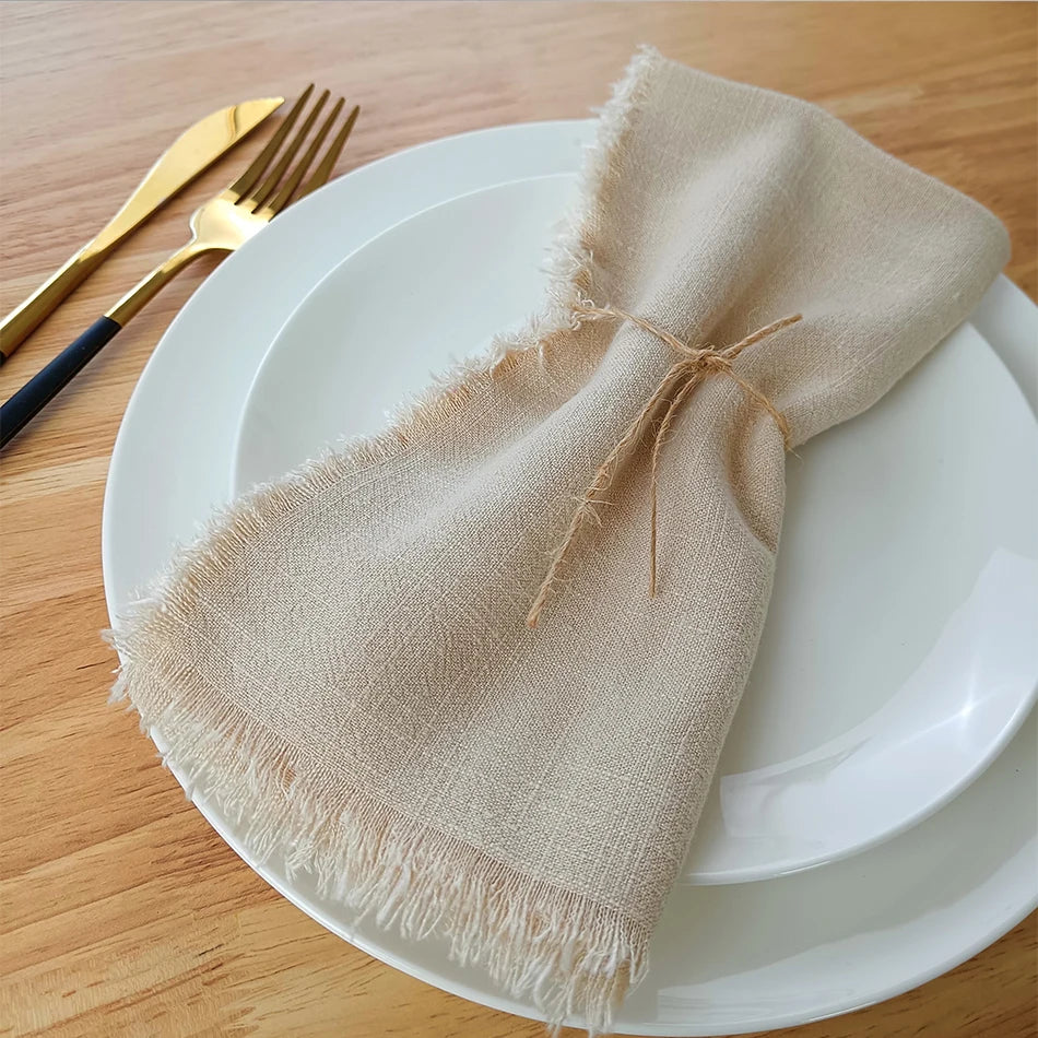 Serviette de table en lin beige, nouée avec une ficelle, placée sur une assiette blanche avec des couverts en métal doré sur une surface en bois.