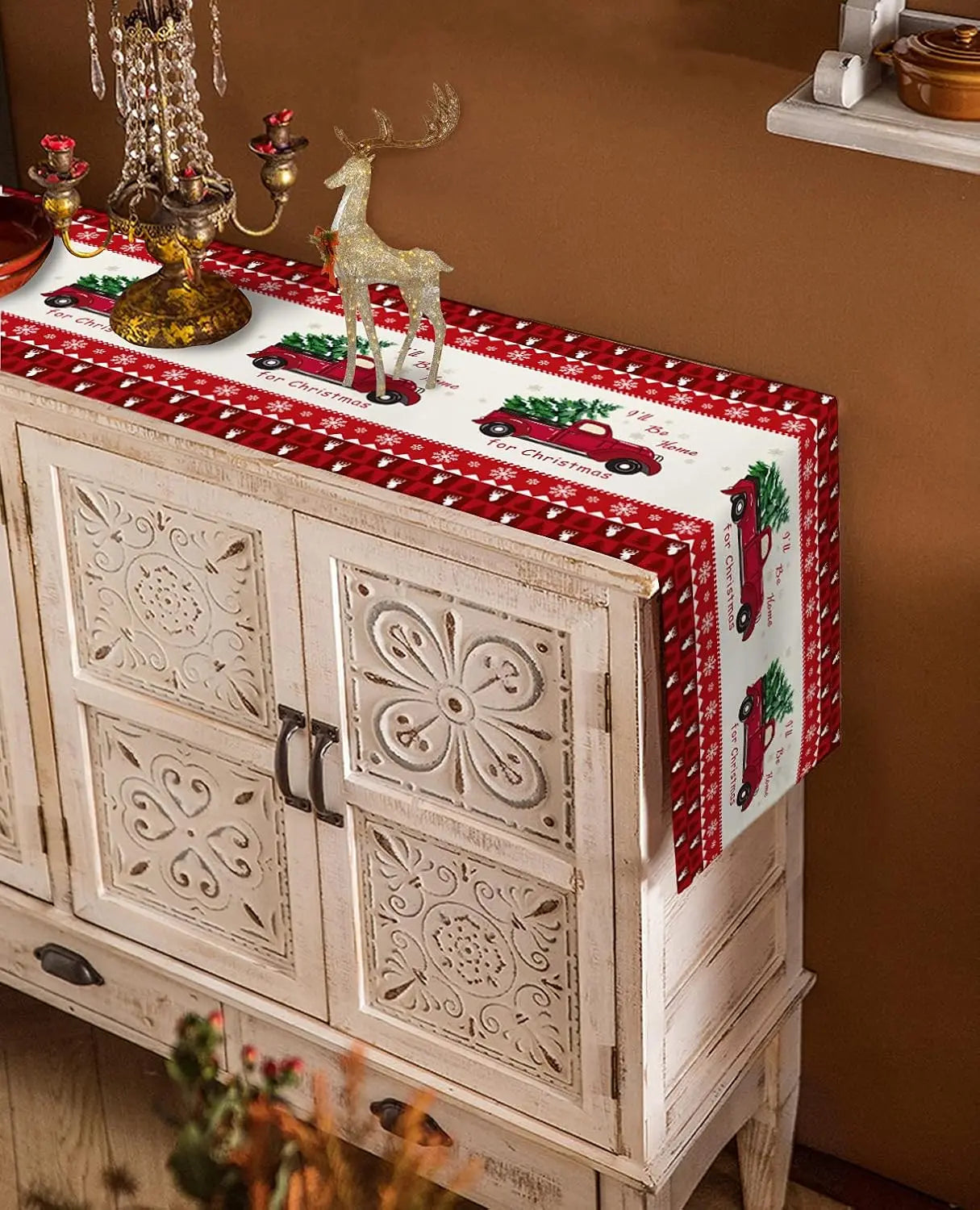 Élégant meuble décoré de motifs sculptés et orné de figurines de Noël sur un élégant chemin de table à imprimé de flocons de neige et de camions rouges, créant une ambiance festive.
