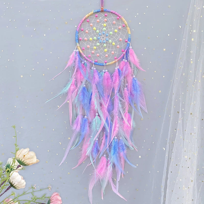 Un attrape-rêves décoratif en forme de spirale, orné de plumes roses, bleues et dorées, avec des perles et des éléments métalliques brillants, suspendu dans un décor avec des fleurs et des étoiles.