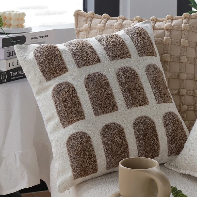 Coussin décoratif beige et marron avec un motif géométrique simple et élégant, posé sur un canapé en osier et accompagné d'un livre et d'une tasse de boisson.