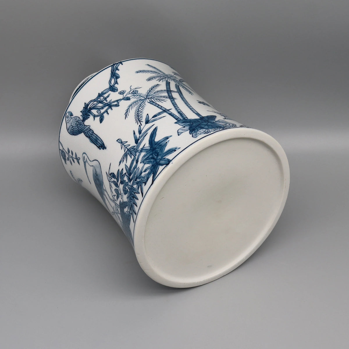 Cette image montre un pot en céramique blanc orné de motifs bleus représentant des feuilles de palmiers, des plantes et des oiseaux.