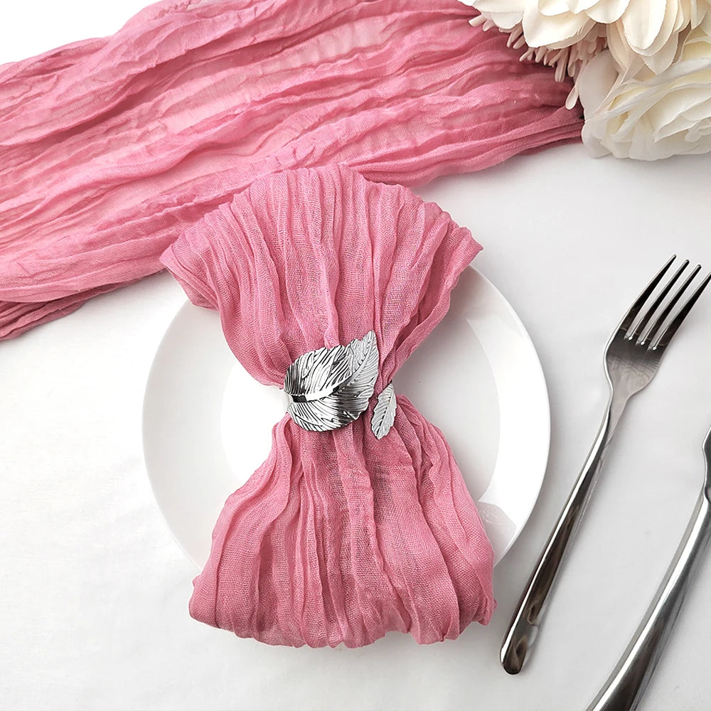 Une serviette de table rose aux plis doux et élégants, avec une décoration en métal en forme de plume, accompagnée d'une fourchette et d'un couteau sur une surface blanche.