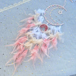 Un attrape-rêves délicat composé de plumes roses et blanches, d'un croissant décoré et d'ornements en perles, sur un fond étoilé bleu.