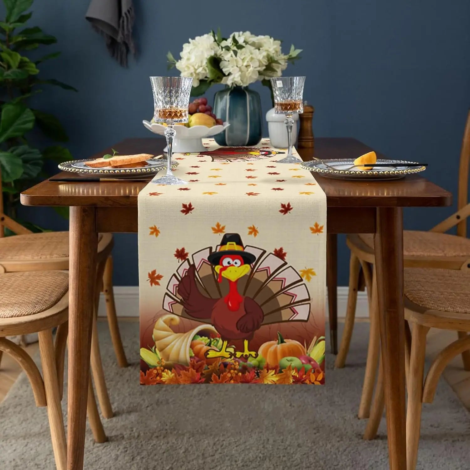 Une jolie table de salle à manger avec une nappe aux motifs d'automne, des verres à vin, des fruits et un bouquet de fleurs blanches, créant une ambiance chaleureuse et festive.