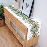 La photo montre un joli meuble en bois clair, sur lequel est posé un chemin de table décoré d'un motif de feuilles et de baies rouges, ainsi qu'un vase blanc et une œuvre d'art abstraite encadrée accrochée au mur.