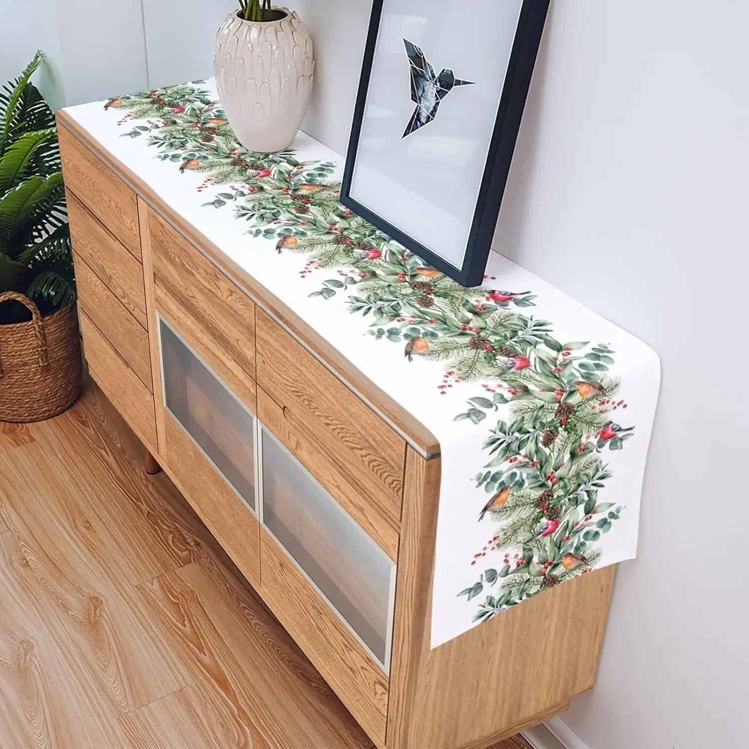 La photo montre un joli meuble en bois clair, sur lequel est posé un chemin de table décoré d'un motif de feuilles et de baies rouges, ainsi qu'un vase blanc et une œuvre d'art abstraite encadrée accrochée au mur.