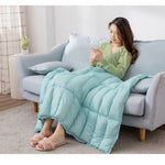 Une jeune femme confortable, en robe verte, enveloppée dans une couverture turquoise, relaxe sur un canapé gris dans un intérieur cosy.