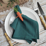 Serviette pliée de couleur verte avec un motif de carotte, placée sur une assiette blanche avec des couverts en bois sur une table en bois.