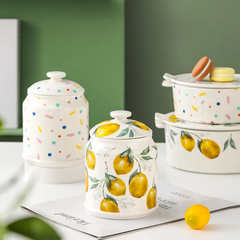 Pot en céramique peint de motifs colorés et de citrons jaunes lumineux, accompagné d'autres récipients assortis, sur un fond blanc et vert, créant une ambiance de cuisine fraîche et ensoleillée.