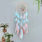 Un capteur de rêves décoratif en macramé avec des plumes roses et bleues, suspendu dans un décor végétal.