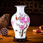 Un magnifique vase en porcelaine blanche orné de délicates fleurs de cerisier roses et d'un oiseau rouge, posé sur un support en bois foncé, créant une atmosphère élégante et apaisante.