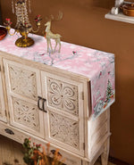 Élégant meuble en bois blanc avec des motifs sculptés, décoré de bougies et d'un cerf doré, sur un fond rose et blanc évoquant une scène hivernale festive.