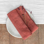 Serviette en lin de couleur terracotta posée sur une assiette ronde et une surface en bois.