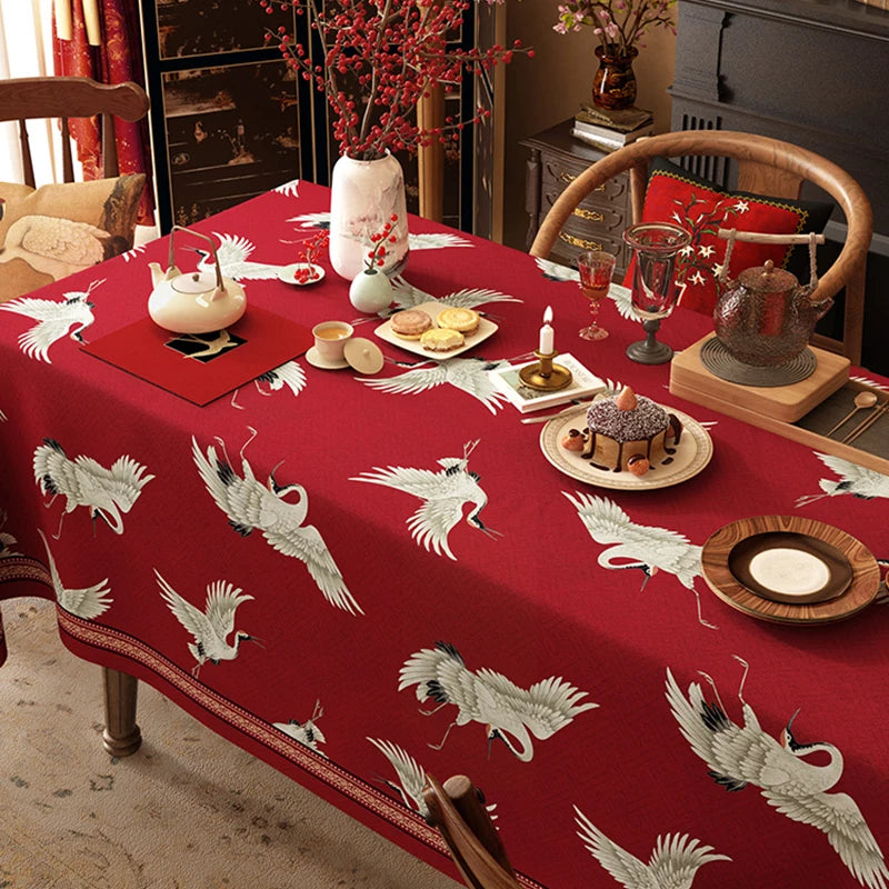 Une table à dîner élégante et festive avec une nappe rouge ornée de grues blanches, une décoration florale, des bougies et des plats traditionnels sur un fond de mobilier vintage.