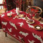 Une table à dîner élégante et festive avec une nappe rouge ornée de grues blanches, une décoration florale, des bougies et des plats traditionnels sur un fond de mobilier vintage.