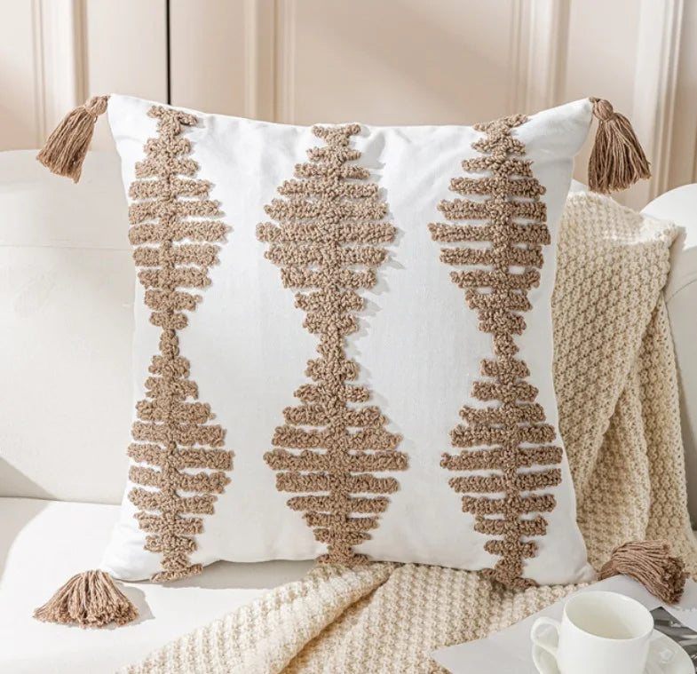 Coussin blanc orné de motifs géométriques texturés en beige, sur fond de couverture en tricot et d'une tasse blanche posées sur un plan de travail clair.