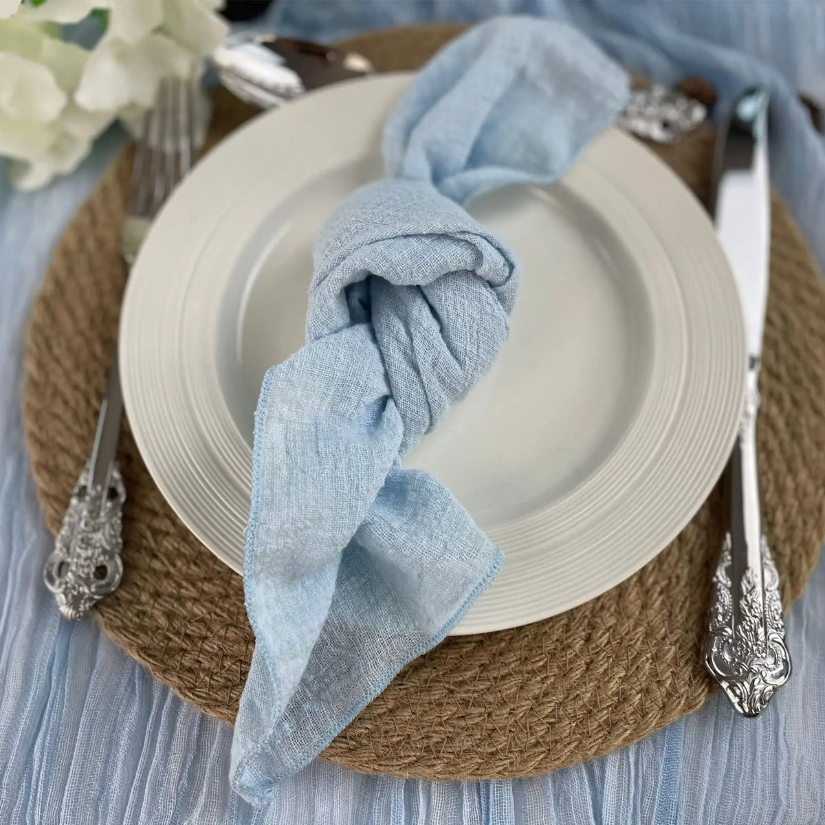 Cette image montre un ensemble de couverts et d'accessoires de table élégants, avec un plat blanc, des ustensiles de qualité et un linge de table bleu gris, sur un fond en fibre naturelle.