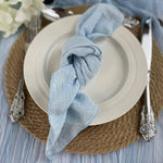 Cette image montre un ensemble de couverts et d'accessoires de table élégants, avec un plat blanc, des ustensiles de qualité et un linge de table bleu gris, sur un fond en fibre naturelle.